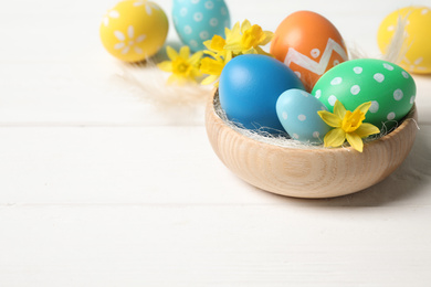 Colorful Easter eggs and flowers in bowl on white wooden table. Space for text