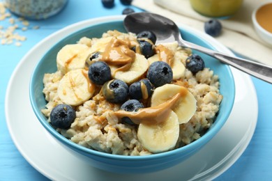 Tasty oatmeal with banana, blueberries and peanut butter served in bowl on light blue wooden table, closeup