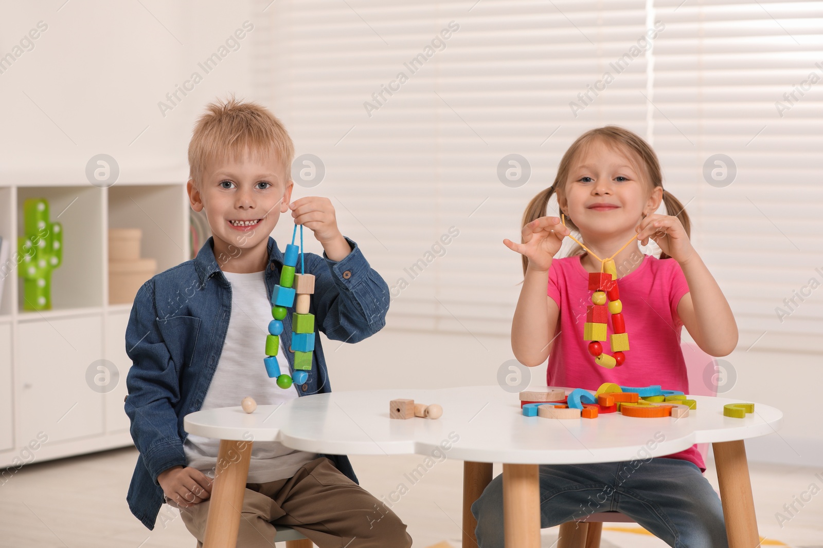 Photo of Little children playing with wooden pieces and string for threading activity at white table indoors. Developmental toys