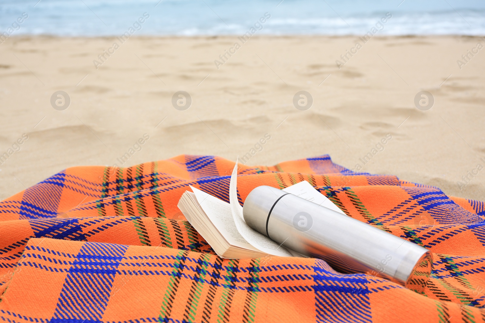 Photo of Metallic thermos with hot drink, open book and plaid on sandy beach near sea