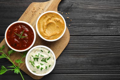 Photo of Different tasty sauces in bowls and parsley on black wooden table, top view. Space for text