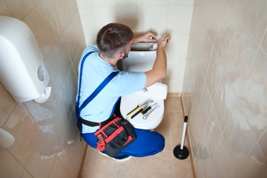 Professional plumber in uniform repairing toilet tank indoors