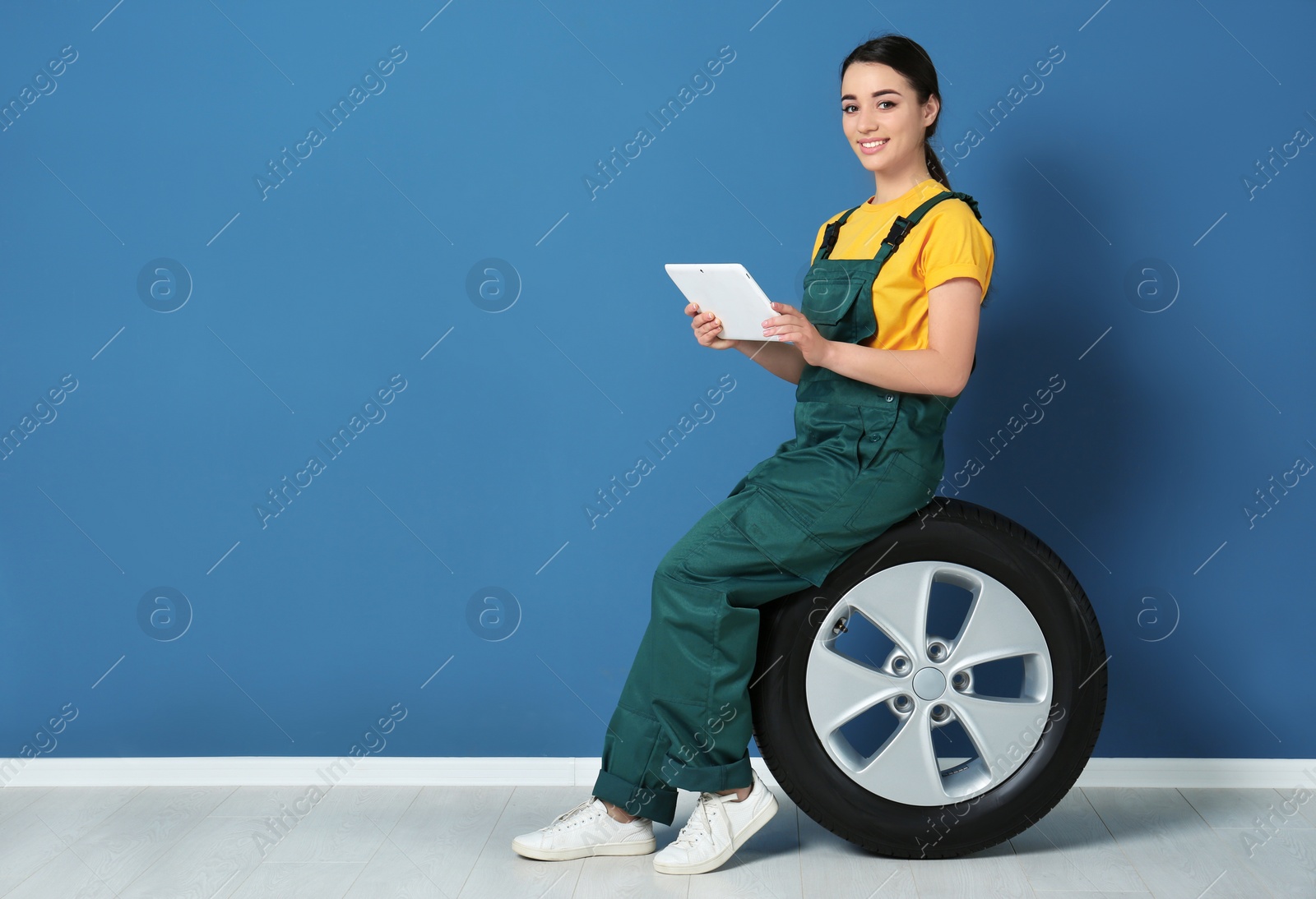 Photo of Female mechanic in uniform with car tire and tablet computer on color wall background