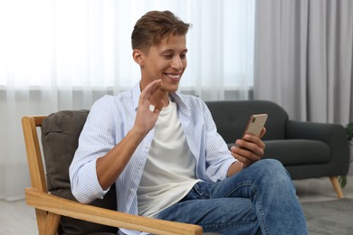Happy young man having video chat via smartphone on armchair indoors