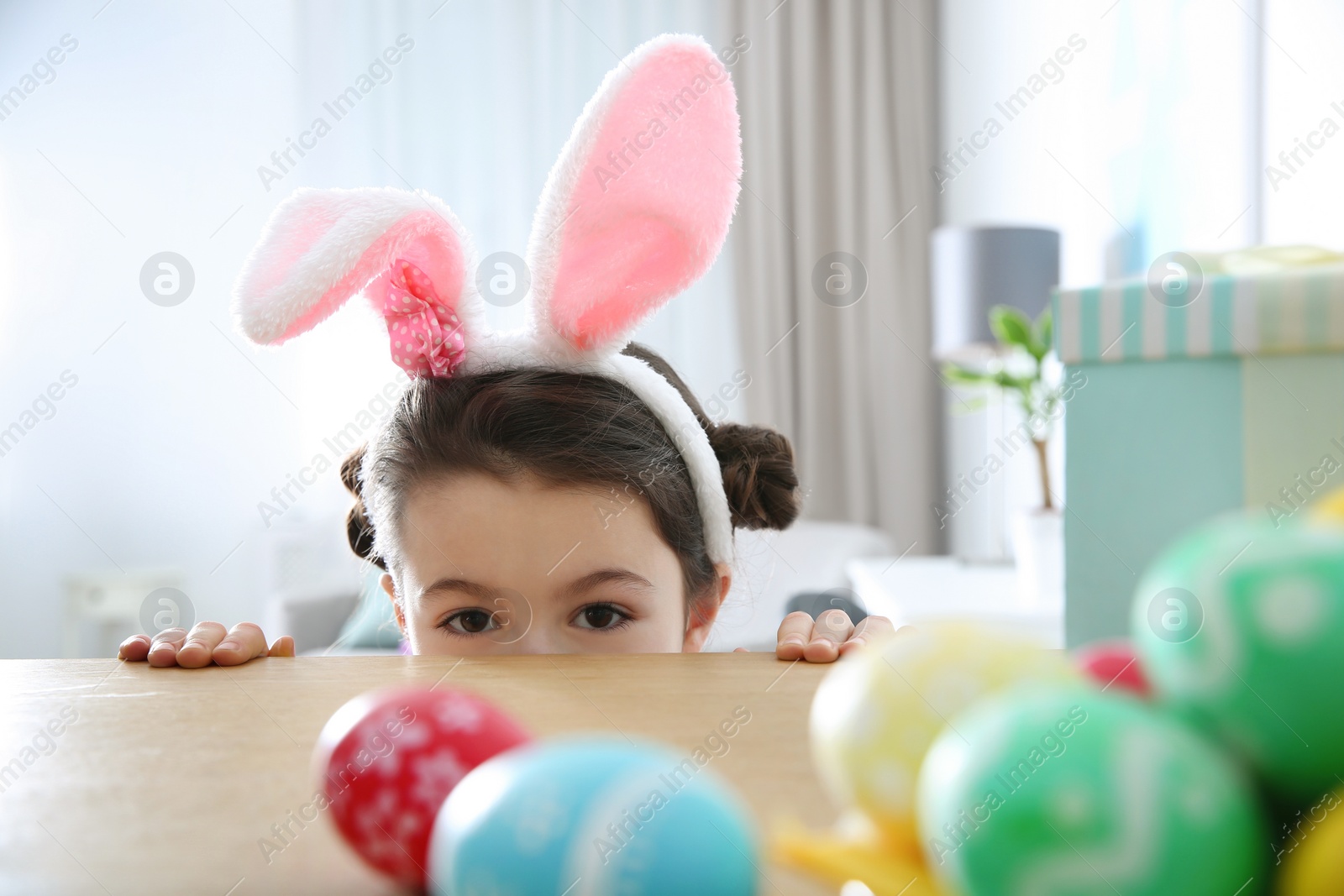 Photo of Cute little girl wearing bunny ears headband hiding behind table with painted Easter eggs in room