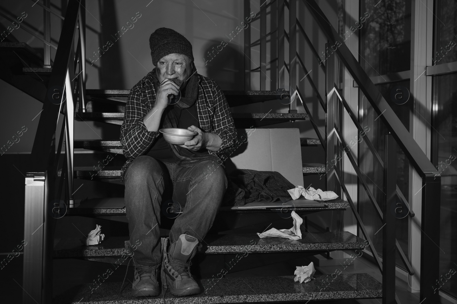 Photo of Poor senior man with bowl and bread on stairs indoors. Black and white effect