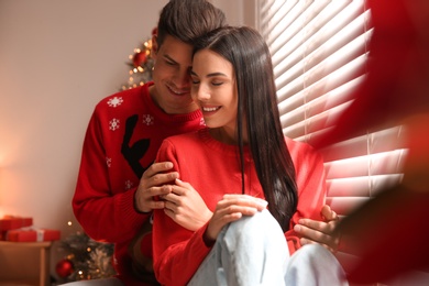 Photo of Happy couple in warm Christmas sweaters near window indoors