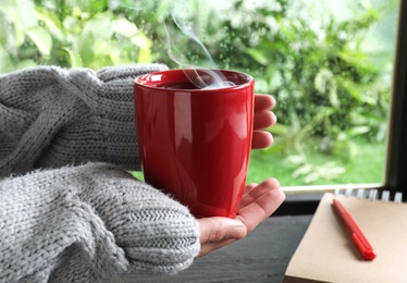 Photo of Woman with cup of hot drink near window on rainy day, closeup