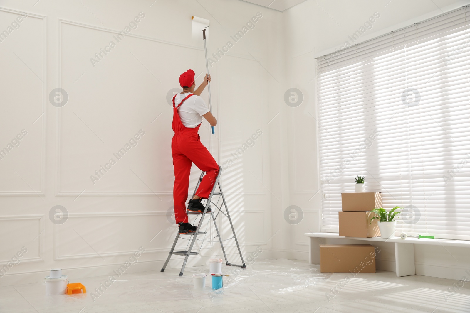 Photo of Worker painting wall with roller on ladder indoors