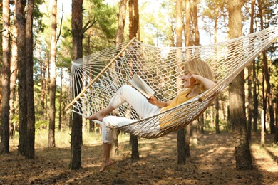 Happy young woman with book relaxing in hammock outdoors on summer day