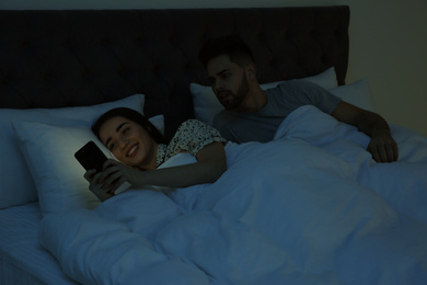 Photo of Distrustful young man peering into girlfriend's smartphone in bed at night