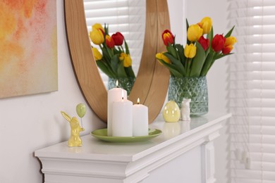 Easter decorations. Bouquet of tulips in vase, burning candles and bunny figures on fireplace at home