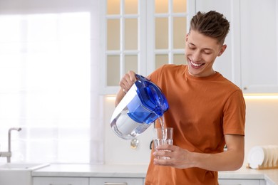 Photo of Happy man pouring water from filter jug into glass in kitchen. Space for text