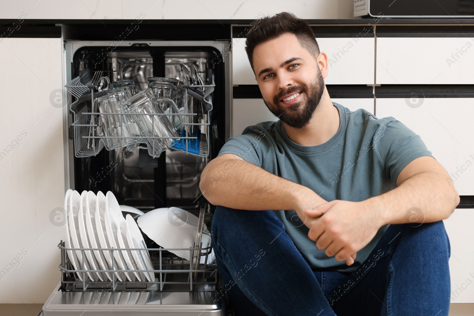 Photo of Smiling man sitting near open dishwasher in kitchen