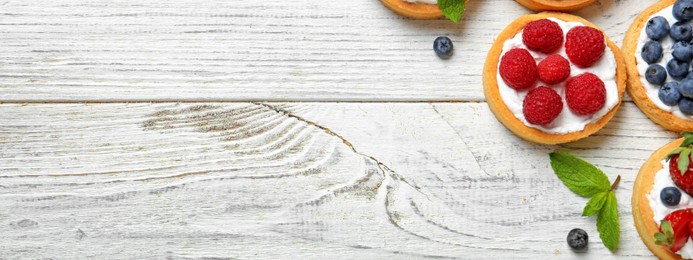 Image of Delicious tartlets with fresh berries on white wooden table, flat lay with space for text. Banner design