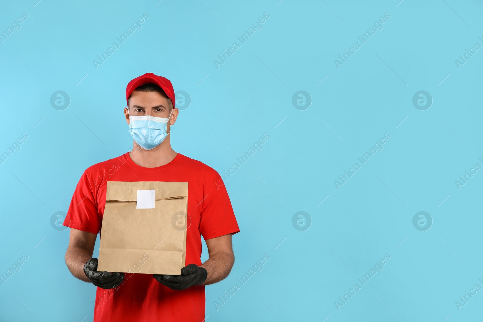 Photo of Courier in medical mask holding paper bag with takeaway food on light blue background, space for text. Delivery service during quarantine due to Covid-19 outbreak
