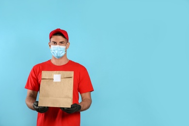 Photo of Courier in medical mask holding paper bag with takeaway food on light blue background, space for text. Delivery service during quarantine due to Covid-19 outbreak