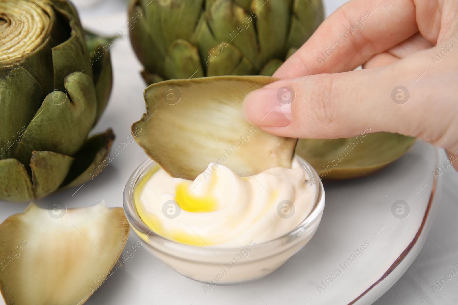 Photo of Woman dipping delicious cooked artichoke into sauce at table, closeup