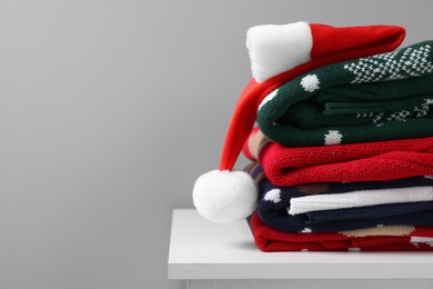 Photo of Different folded Christmas sweaters and Santa hat on chest of drawers against grey background. Space for text