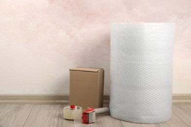 Photo of Bubble wrap roll, tape dispenser and cardboard box on floor near pink wall. Space for text