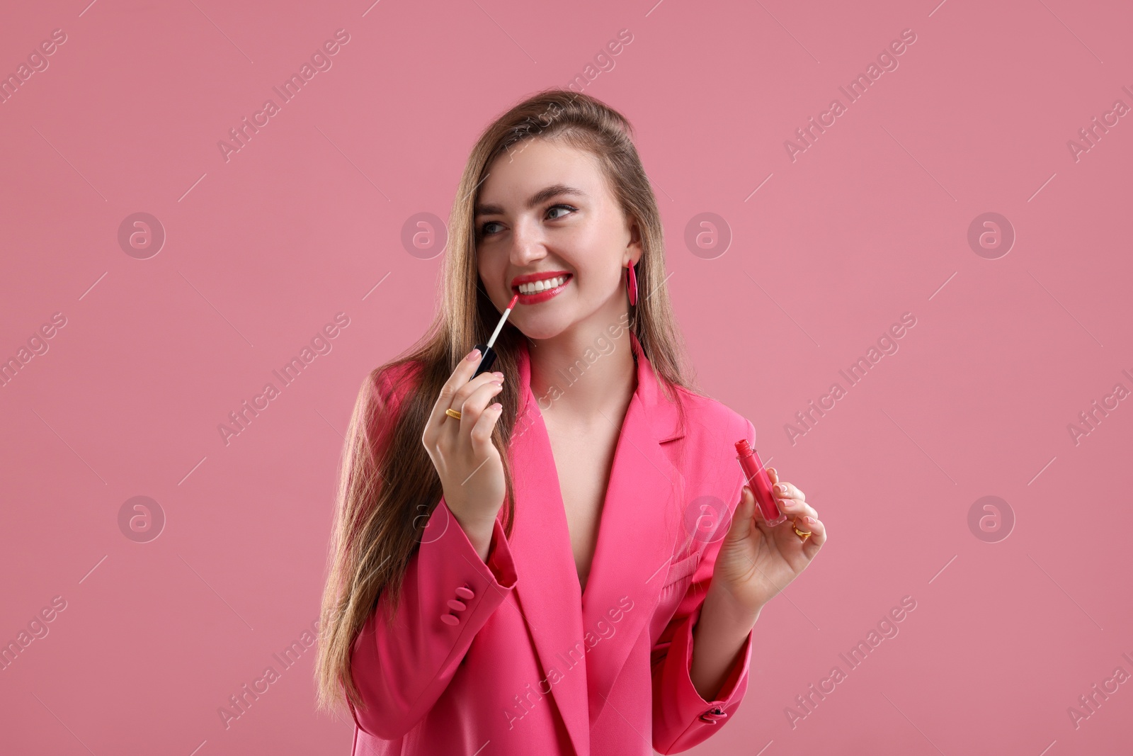 Photo of Beautiful woman in pink clothes applying lip gloss against color background