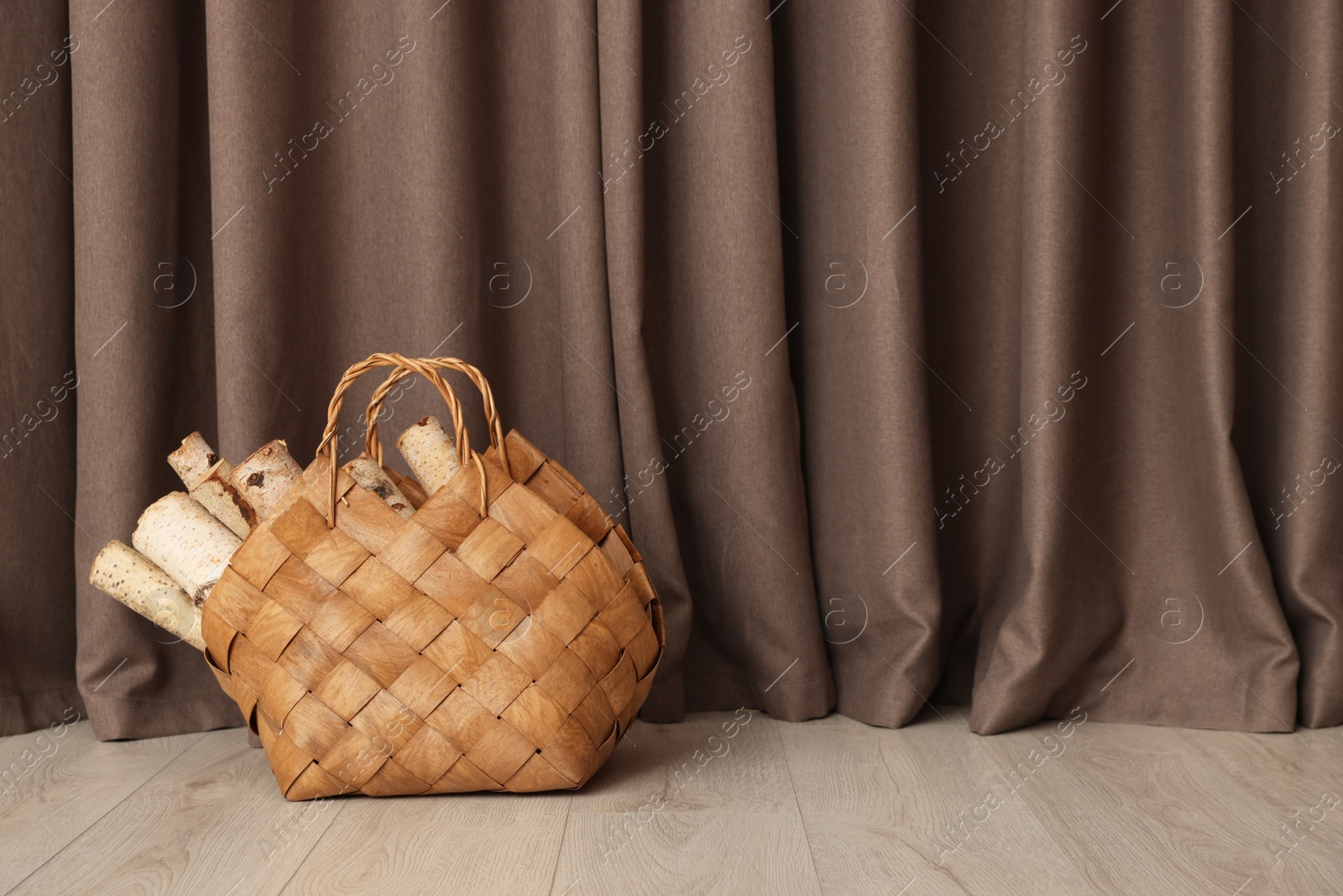 Photo of Wicker bag with firewood on floor near elegant curtains indoors, space for text