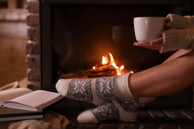 Photo of Woman in knitted socks holding hot drink near fireplace at home, closeup. Winter vacation