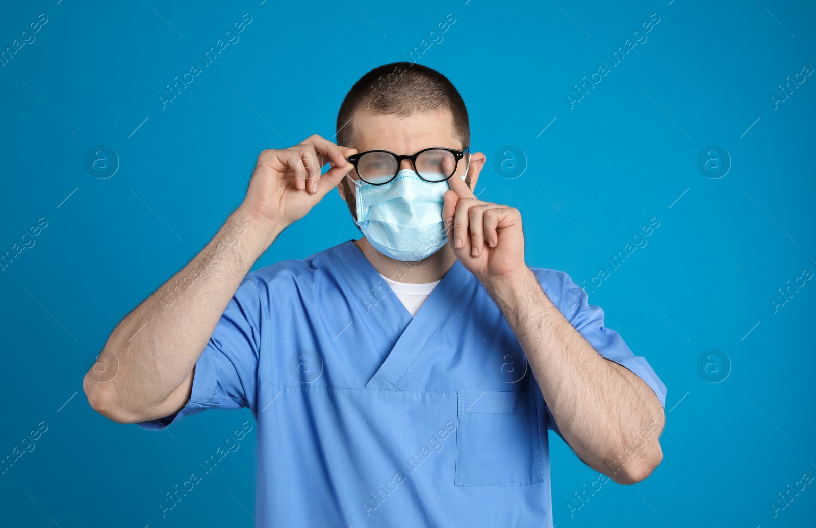 Photo of Doctor wiping foggy glasses caused by wearing disposable mask on blue background. Protective measure during coronavirus pandemic