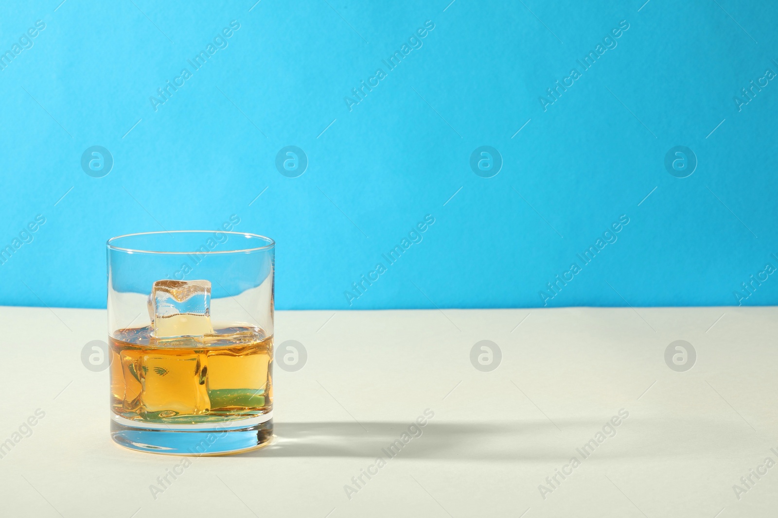 Photo of Whiskey with ice cubes in glass on white table against light blue background, space for text