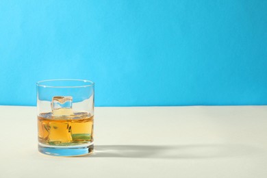 Photo of Whiskey with ice cubes in glass on white table against light blue background, space for text