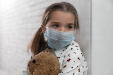Sad little girl in protective mask looking out of window indoors, view from outside. Staying at home during coronavirus pandemic