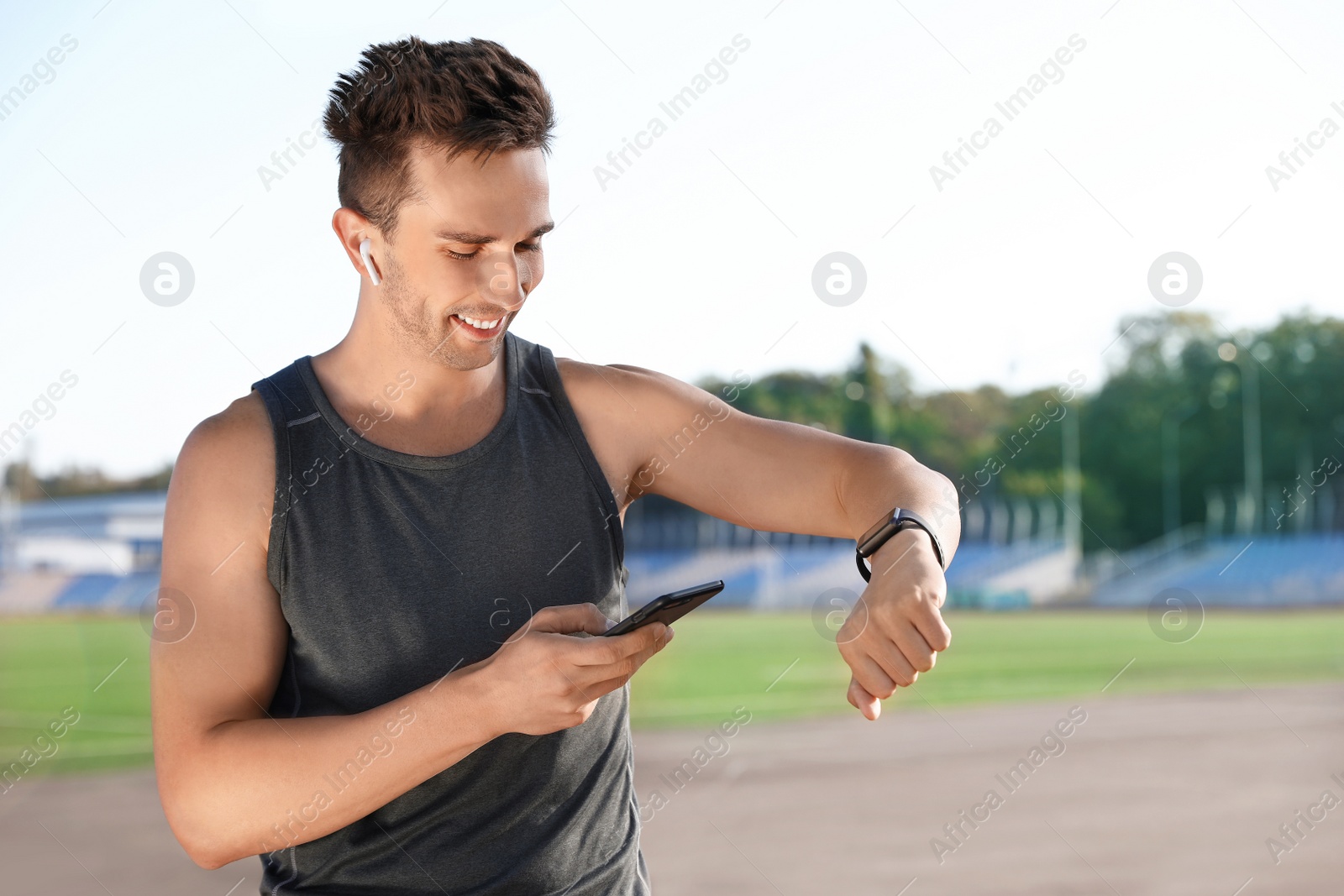 Photo of Young sportsman with wireless earphones, mobile phone and smart watch at stadium