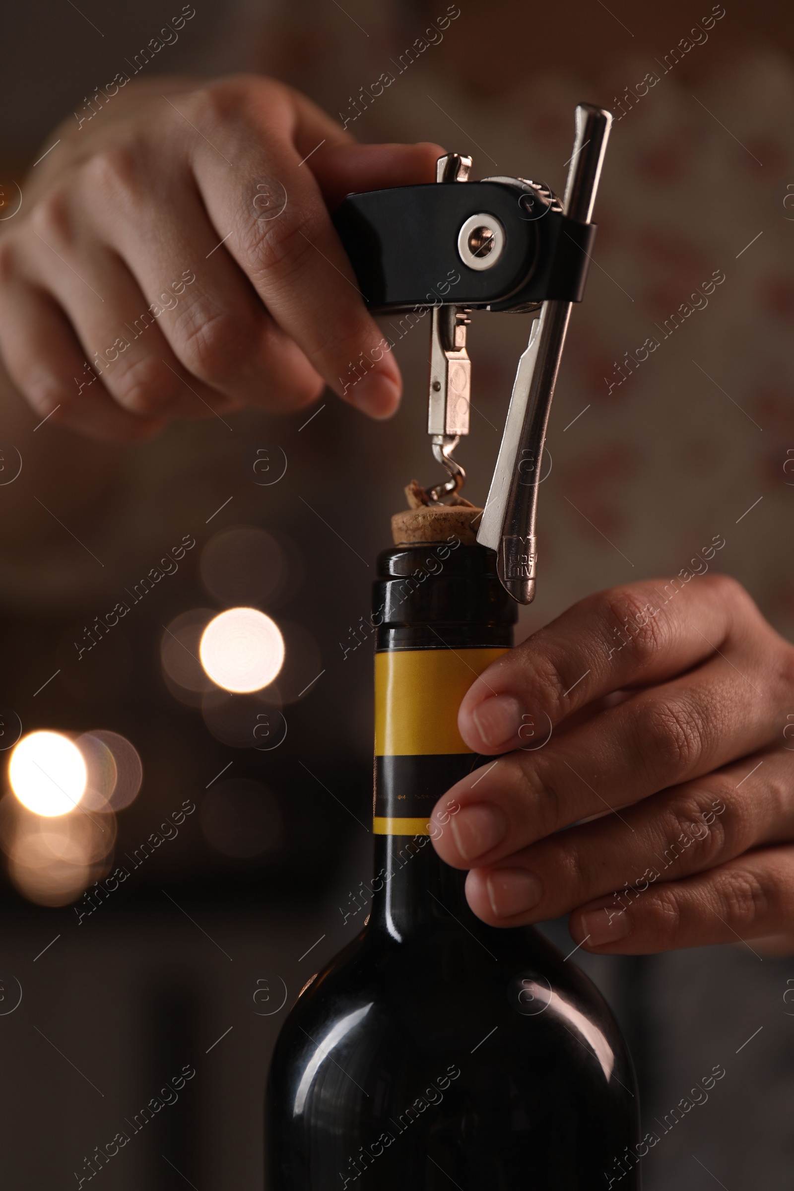 Photo of Woman opening wine bottle with corkscrew on blurred background, closeup