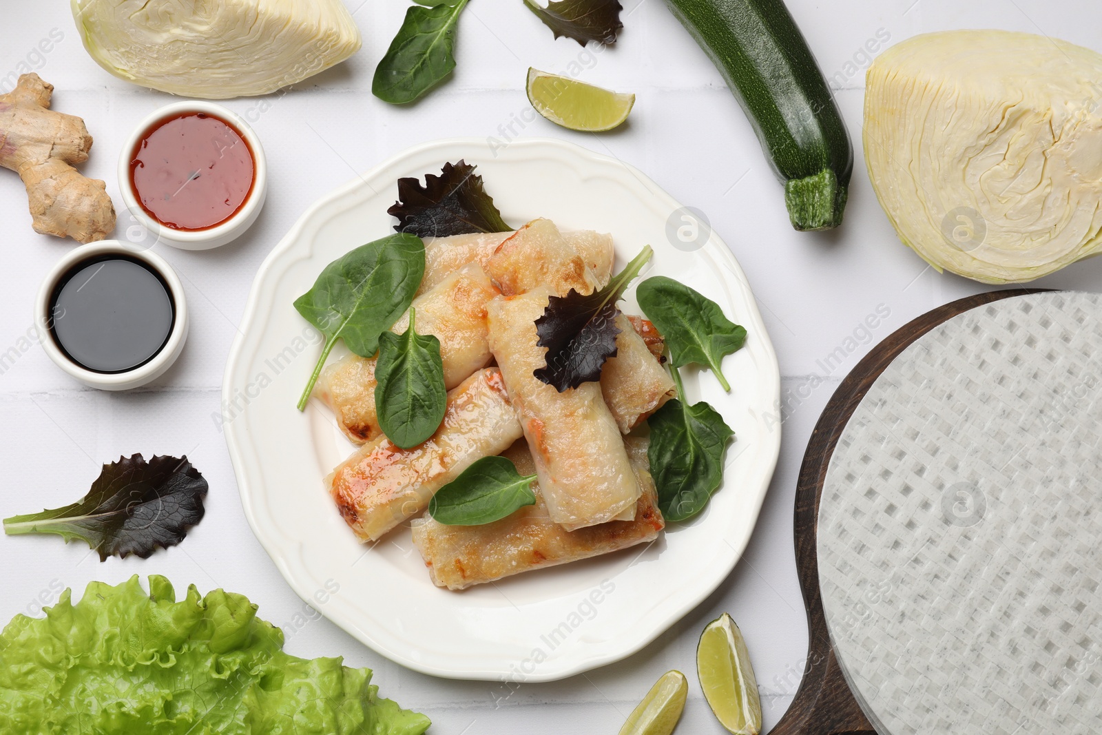 Photo of Plate with tasty fried spring rolls, spinach, sauces and other products on white tiled table, flat lay