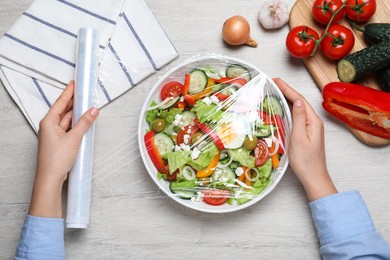 Photo of Woman putting plastic food wrap over bowl of fresh salad at wooden table, top view