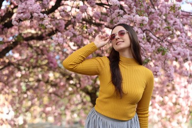 Beautiful woman in sunglasses near blossoming tree on spring day, space for text