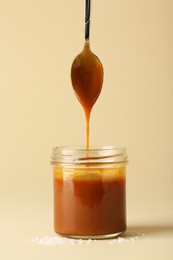 Photo of Pouring tasty salted caramel from spoon into jar on pale yellow background