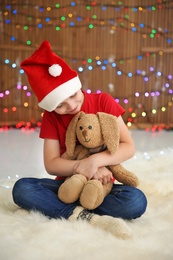 Photo of Cute little child in Santa hat with toy rabbit on blurred lights background. Christmas celebration