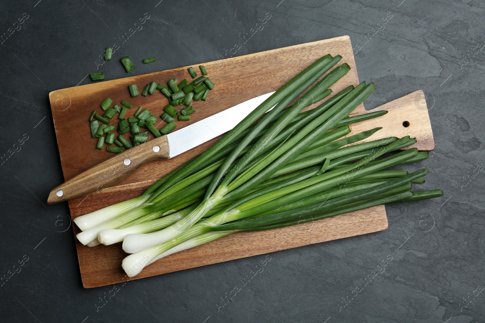 Photo of Fresh green spring onions, cutting board and knife on black table, top view