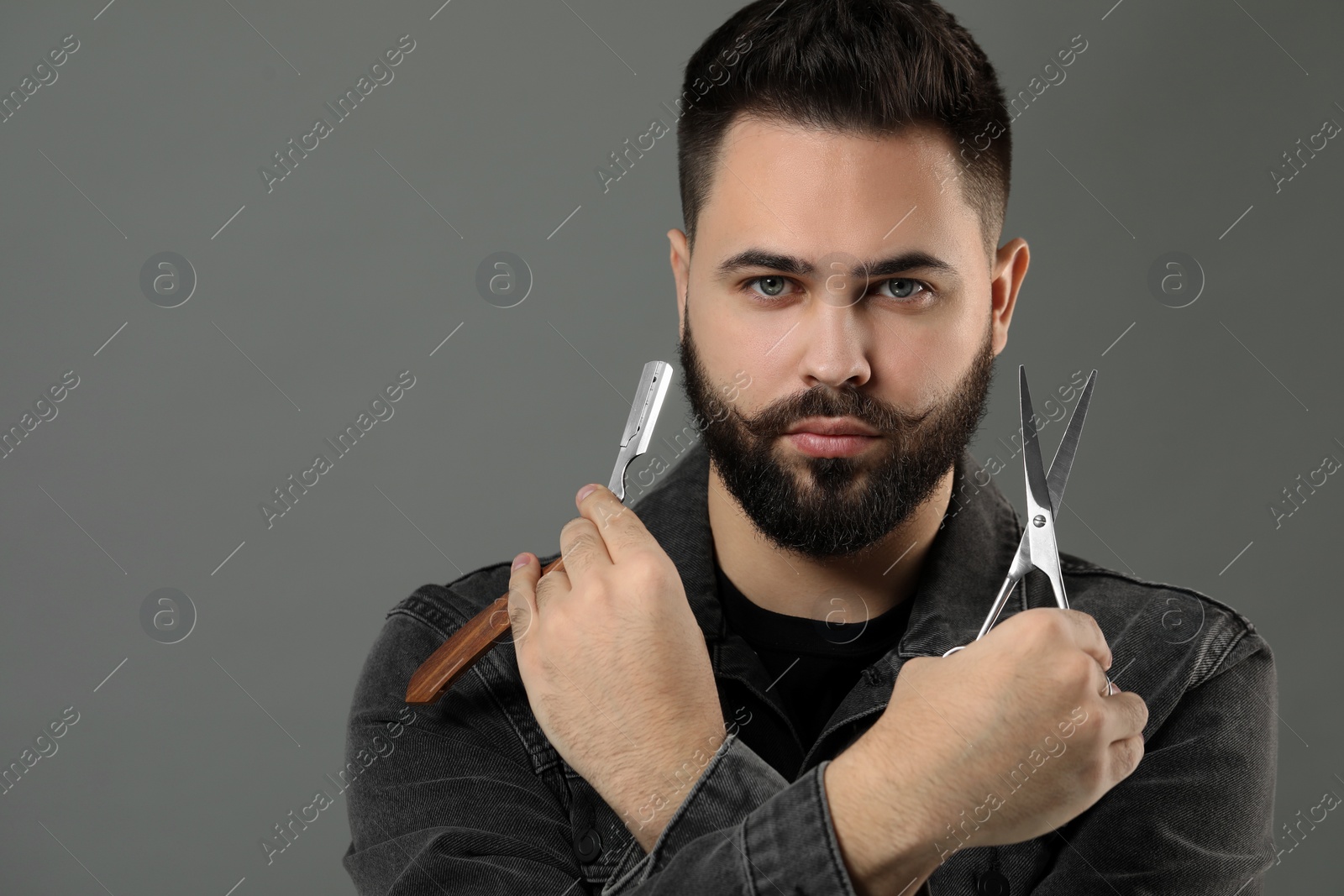 Photo of Handsome young man with mustache holding blade and scissors on grey background