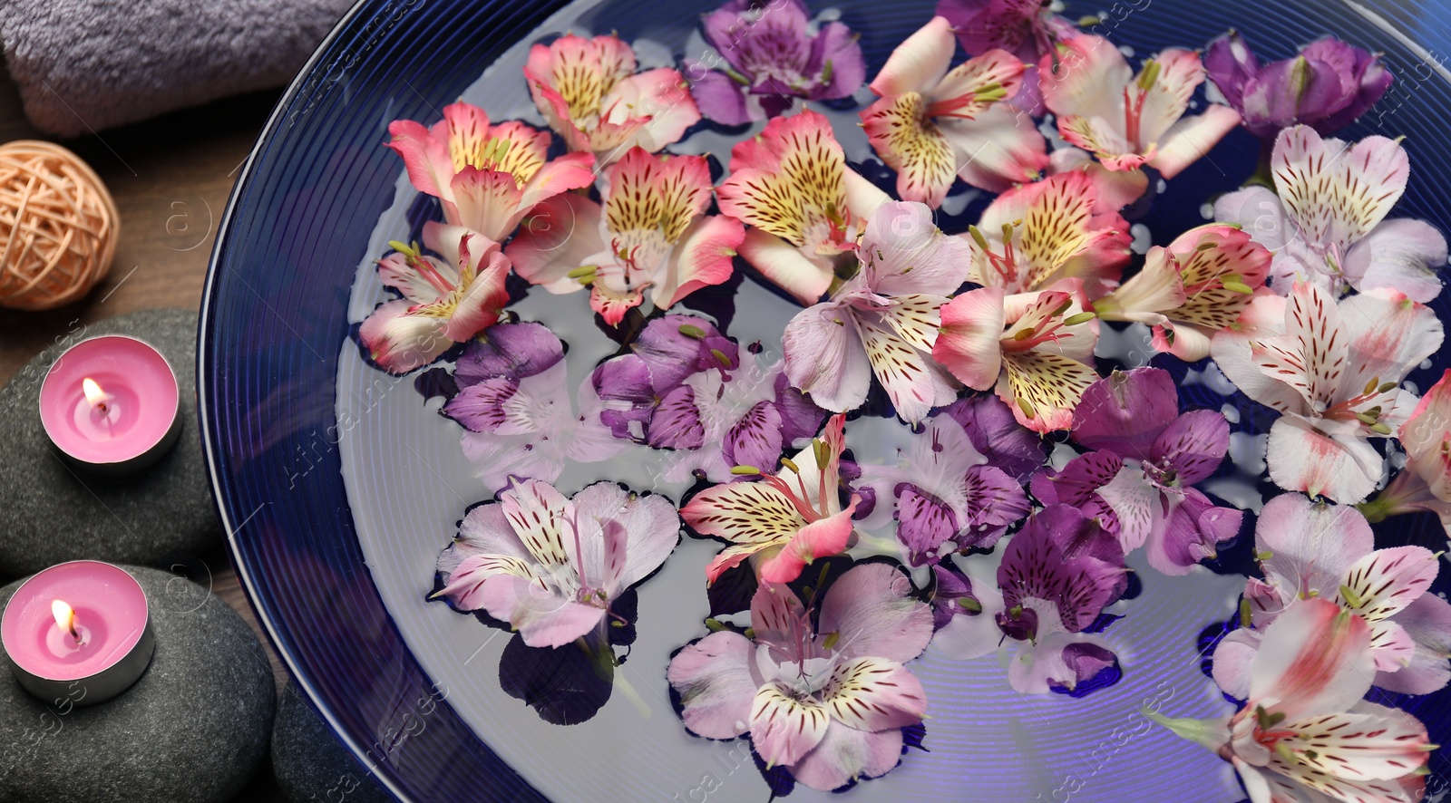 Photo of Bowl of water with flowers, candles and stones on table, closeup. Spa treatment