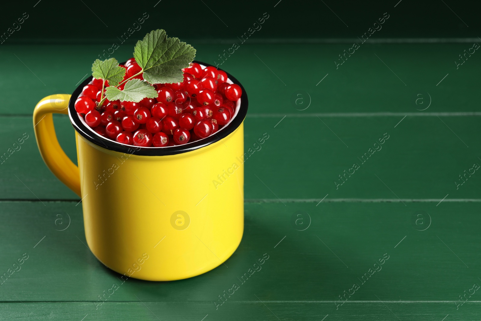 Photo of Ripe red currants and leaves in mug on green wooden table. Space for text