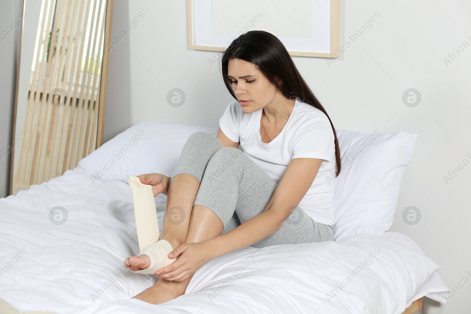 Photo of Young woman applying medical bandage onto foot in bedroom