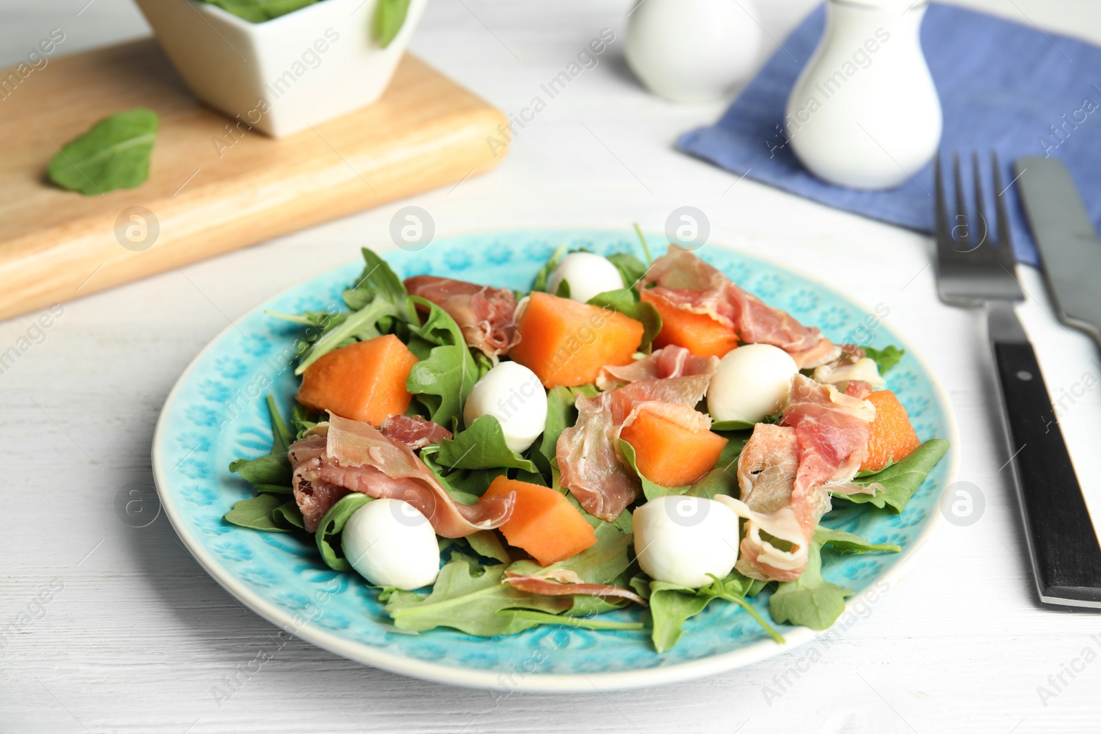 Photo of Fresh melon with prosciutto, mozzarella and arugula on white wooden table