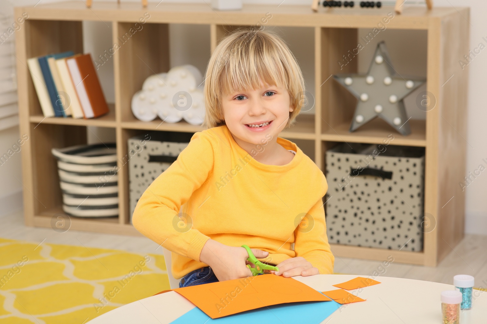 Photo of Cute little boy cutting orange paper at desk in room. Home workplace