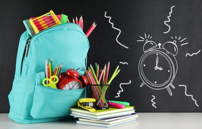 Backpack full of stationery near blackboard with drawn alarm clock. Time to school