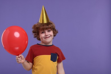 Photo of Happy little boy in party hat with balloon on purple background. Space for text