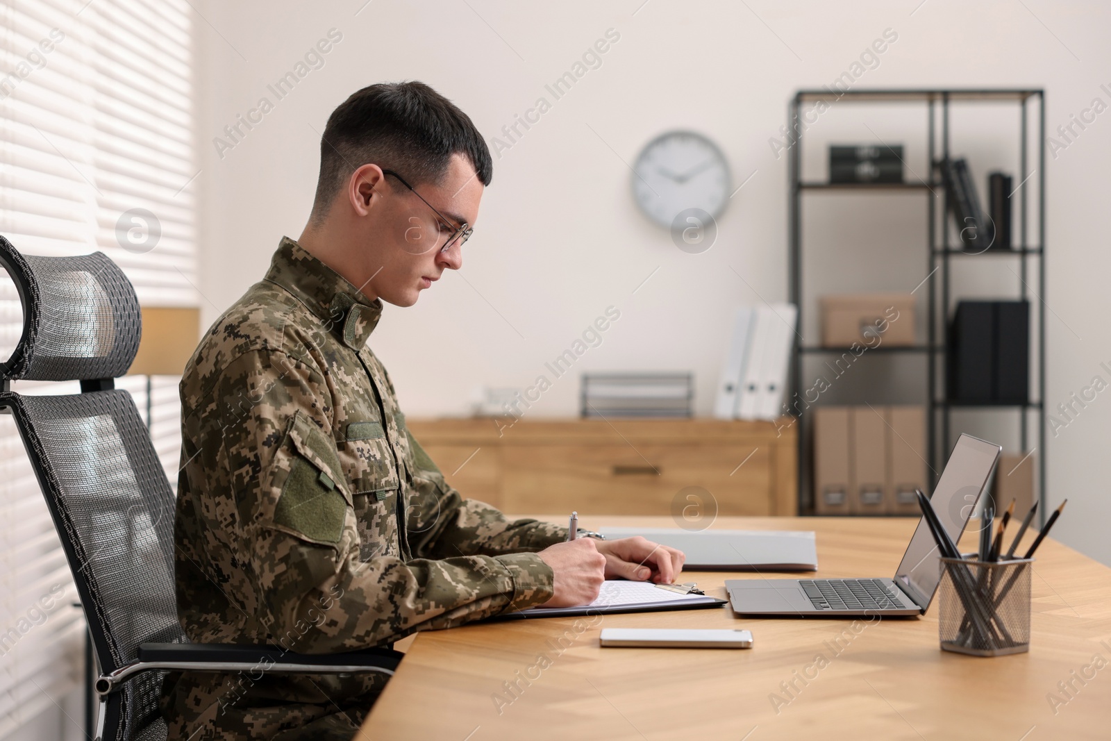 Photo of Military service. Young soldier working at wooden table in office