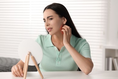 Suffering from allergy. Young woman looking in mirror and scratching her neck at white table indoors