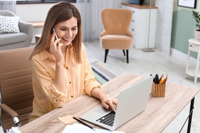 Photo of Young woman talking on phone while working with laptop at desk in home office